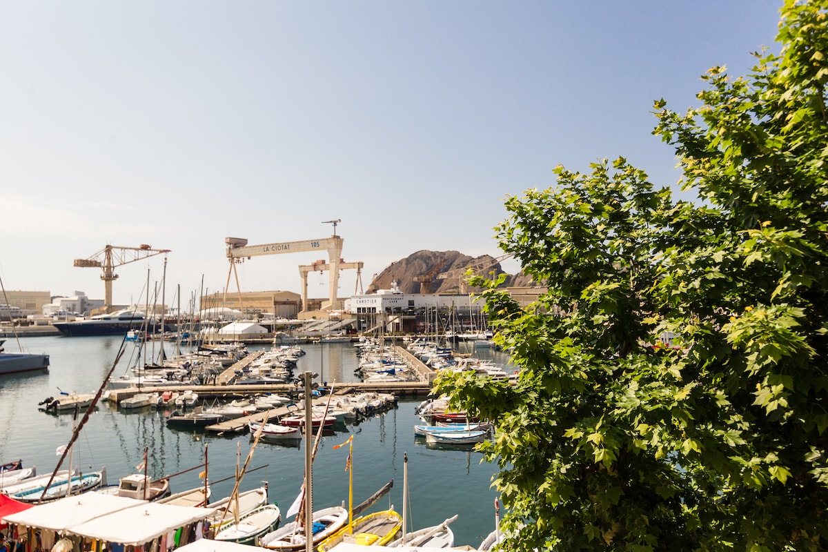 The image depicts a serene view of a marina featuring numerous boats docked in calm waters. A tree is partially visible in the foreground, adding greenery to the scene. In the background, industrial cranes and rugged hills contribute to the port's distinctive atmosphere.