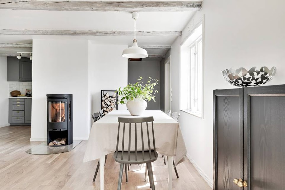 A dining area is presented with a light-colored table surrounded by gray chairs. A modern wood stove is positioned nearby, providing warmth to the space. Natural light pours in through the windows, enhancing the simple, clean design of the room.