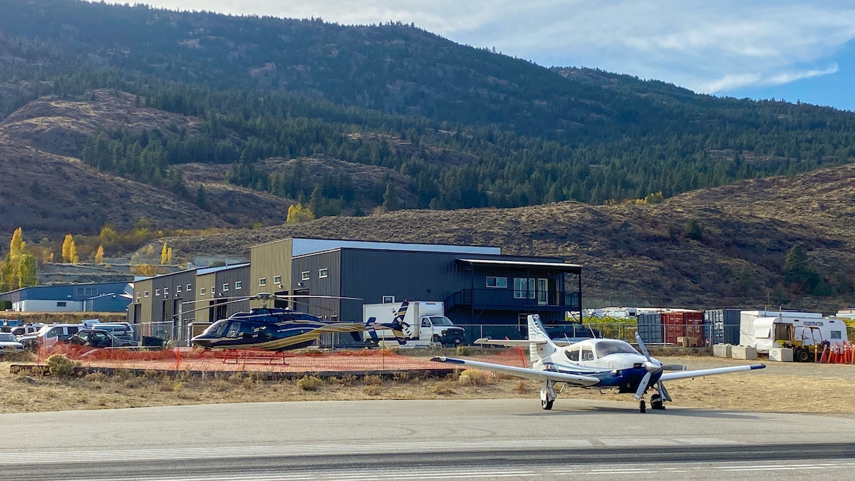A clear view of the upgraded Osoyoos Airport showcases a paved apron with various aircraft parked nearby. The modern terminal building, set against picturesque mountainous terrain, features a facade of dark materials. Surrounding areas include parked vehicles and storage facilities.