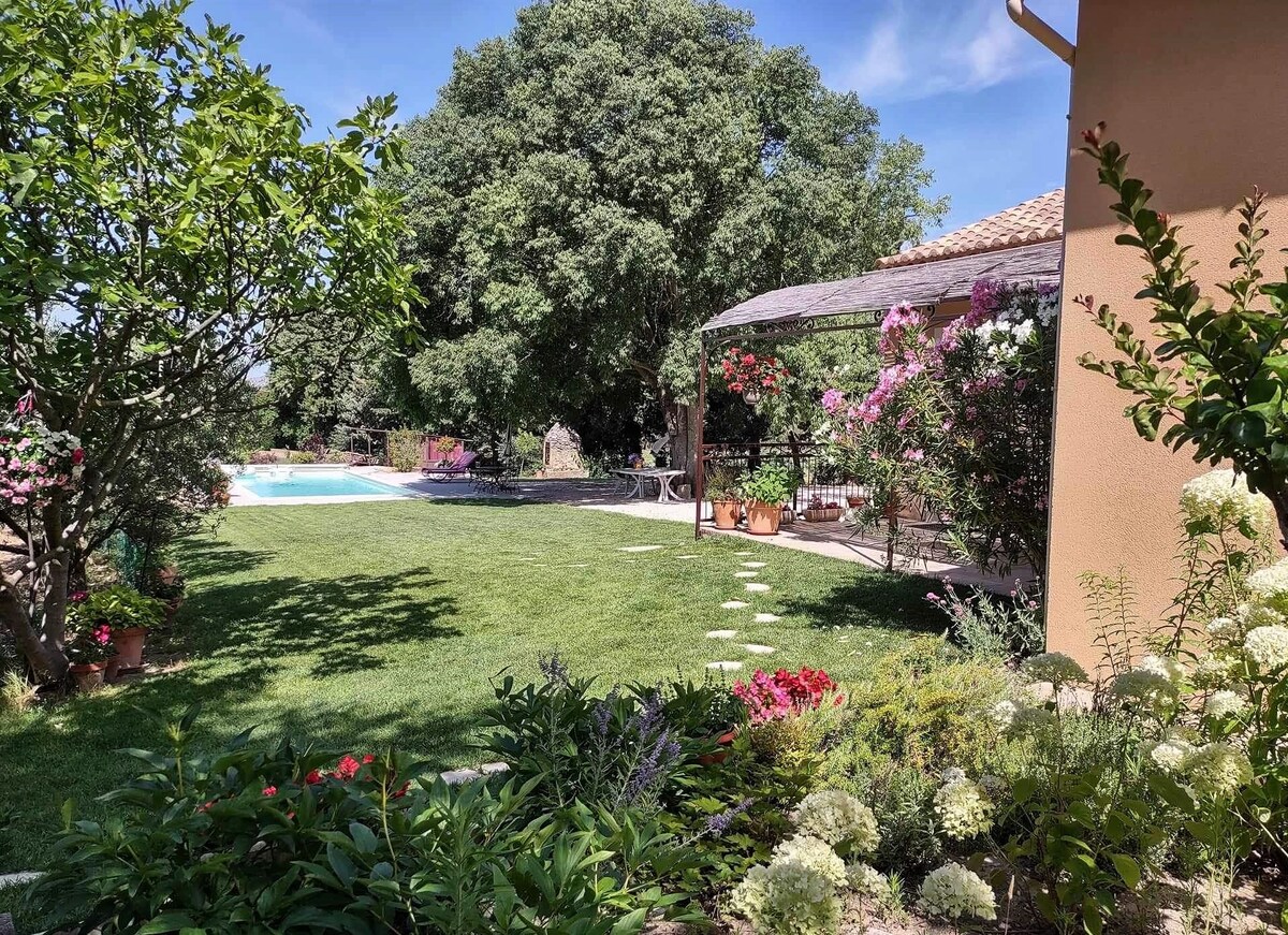 A sunlit garden is featured, showcasing lush green grass and a variety of blooming flowers. In the background, a sparkling pool is visible, surrounded by trees and climbing plants, while a seating area offers a serene space for relaxation.