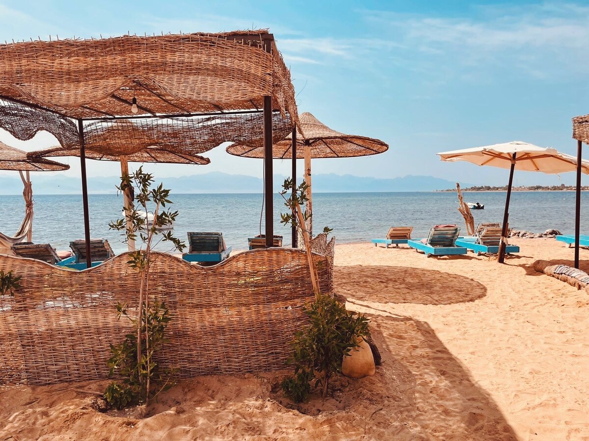 The sandy beach area features woven shade structures providing shelter over lounge chairs. Sun loungers in the background face the calm waters, while light umbrellas stand nearby. Gentle waves are visible in the distance, with a mountain range framed against a clear sky.