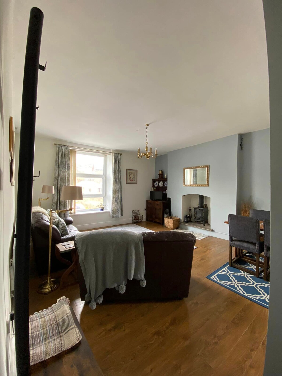 A spacious living area is presented with a comfortable brown sofa covered by a grey throw. Natural light enters through the window, illuminating the wooden floor. A dining table sits to the side, and a decorative fireplace adds warmth to the ambiance.