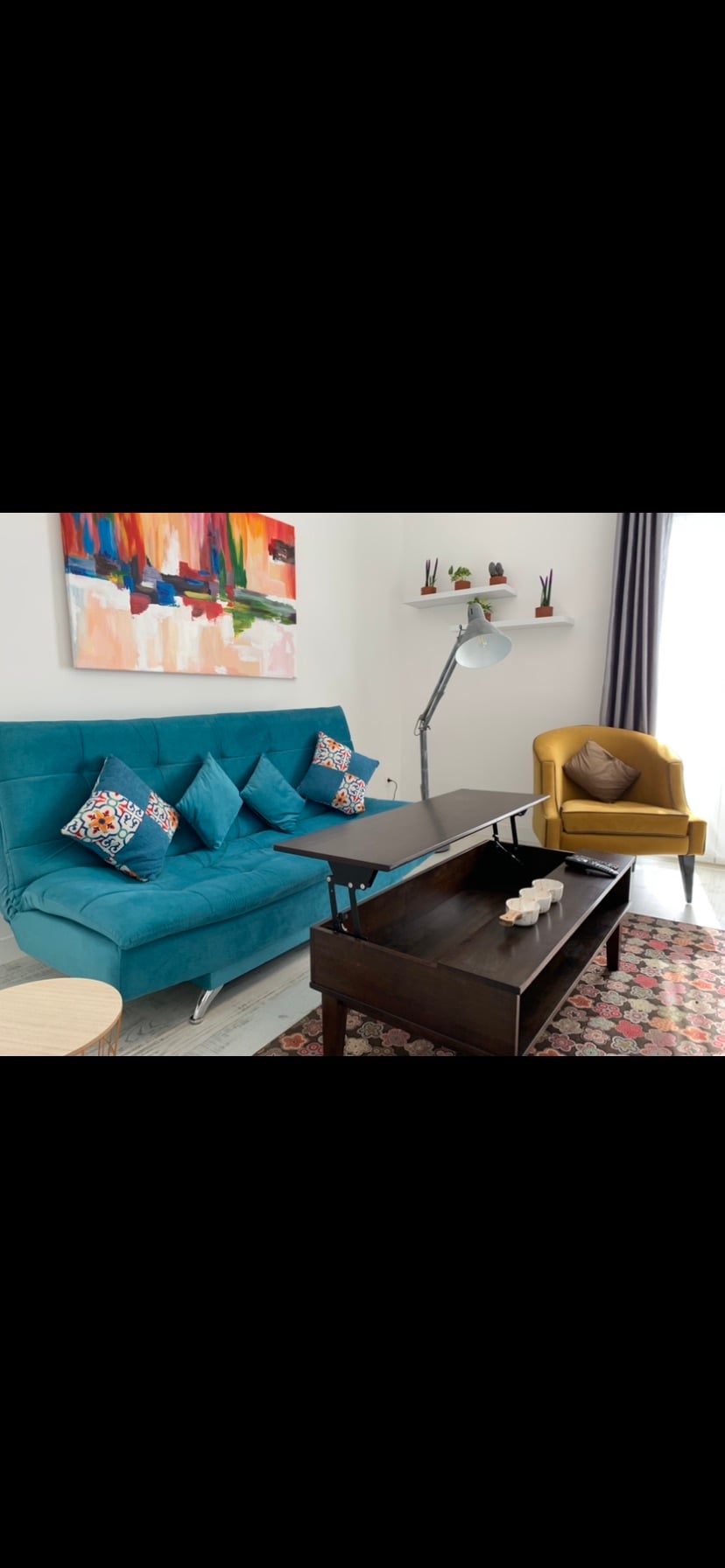 A modern living area features a vibrant blue sofa adorned with patterned cushions. A sleek wooden coffee table is placed in front, with decorative items arranged on top. An armchair in a warm yellow hue complements the space, while a colorful artwork hangs on the wall.