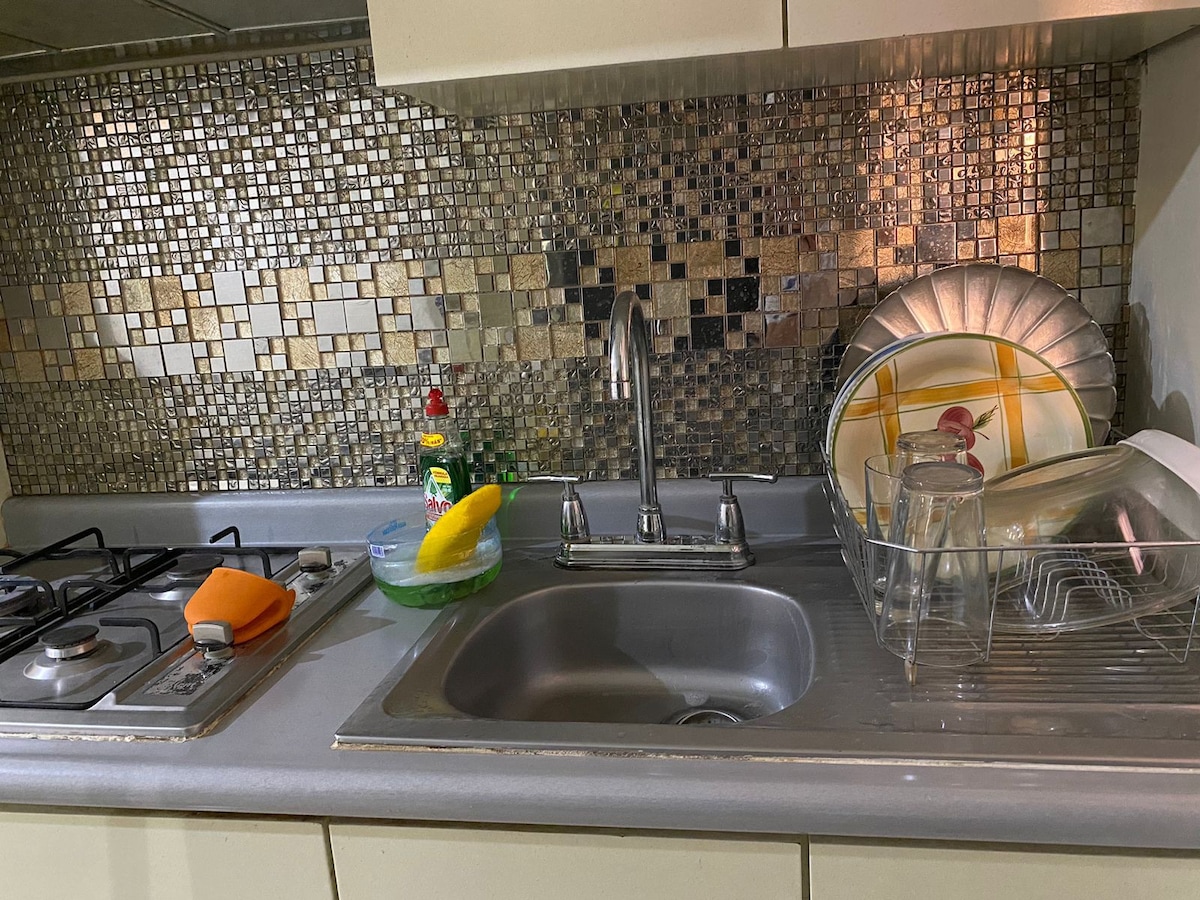 The kitchen area features a stainless steel sink and a tiled backsplash with a mosaic design. A gas stove is positioned alongside dishware, including glasses and a plate. Cleaning supplies are neatly arranged, providing a functional cooking space.