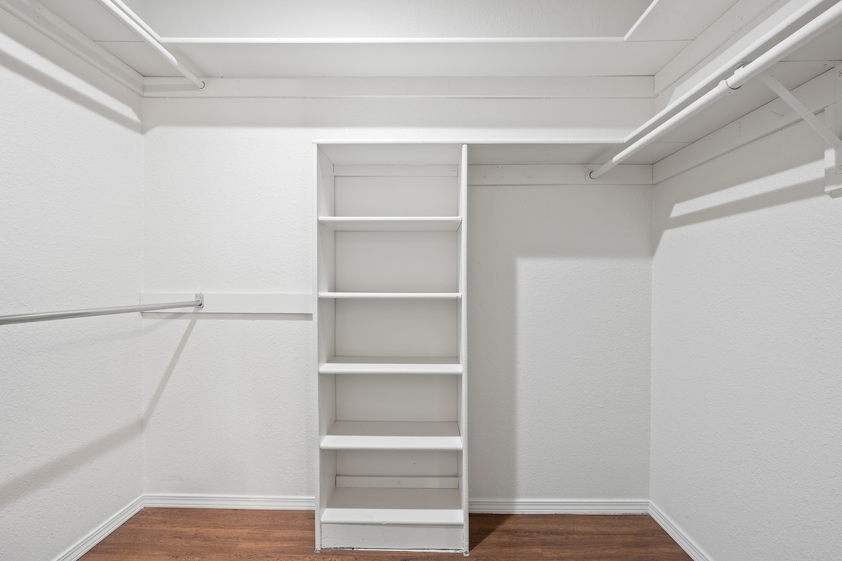 A spacious closet is featured, with light-colored walls and a wooden floor. One empty shelving unit is visible against one wall, while hanging rods are installed along the opposite wall, providing ample space for storage.