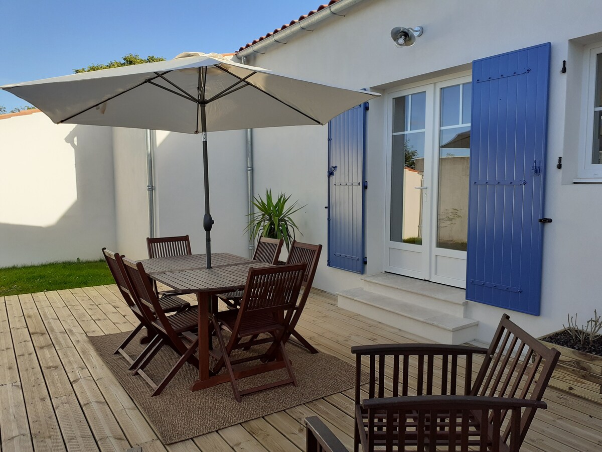 An outdoor seating area is depicted, featuring a large wooden table surrounded by six wooden chairs. A large umbrella provides shade. Two wooden armchairs are positioned on the side, with a plant visible nearby. The light-colored wall and blue shutters create a serene backdrop.