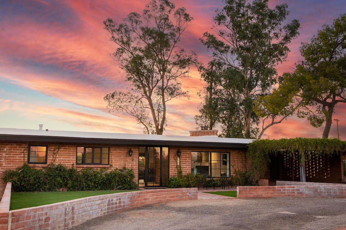 The exterior of a Mid-century home is showcased against a vibrant sunset sky. Large glass doors and windows reflect the colorful clouds. The surrounding landscape features manicured green grass and trees, framing the entrance with a warm and inviting ambiance.