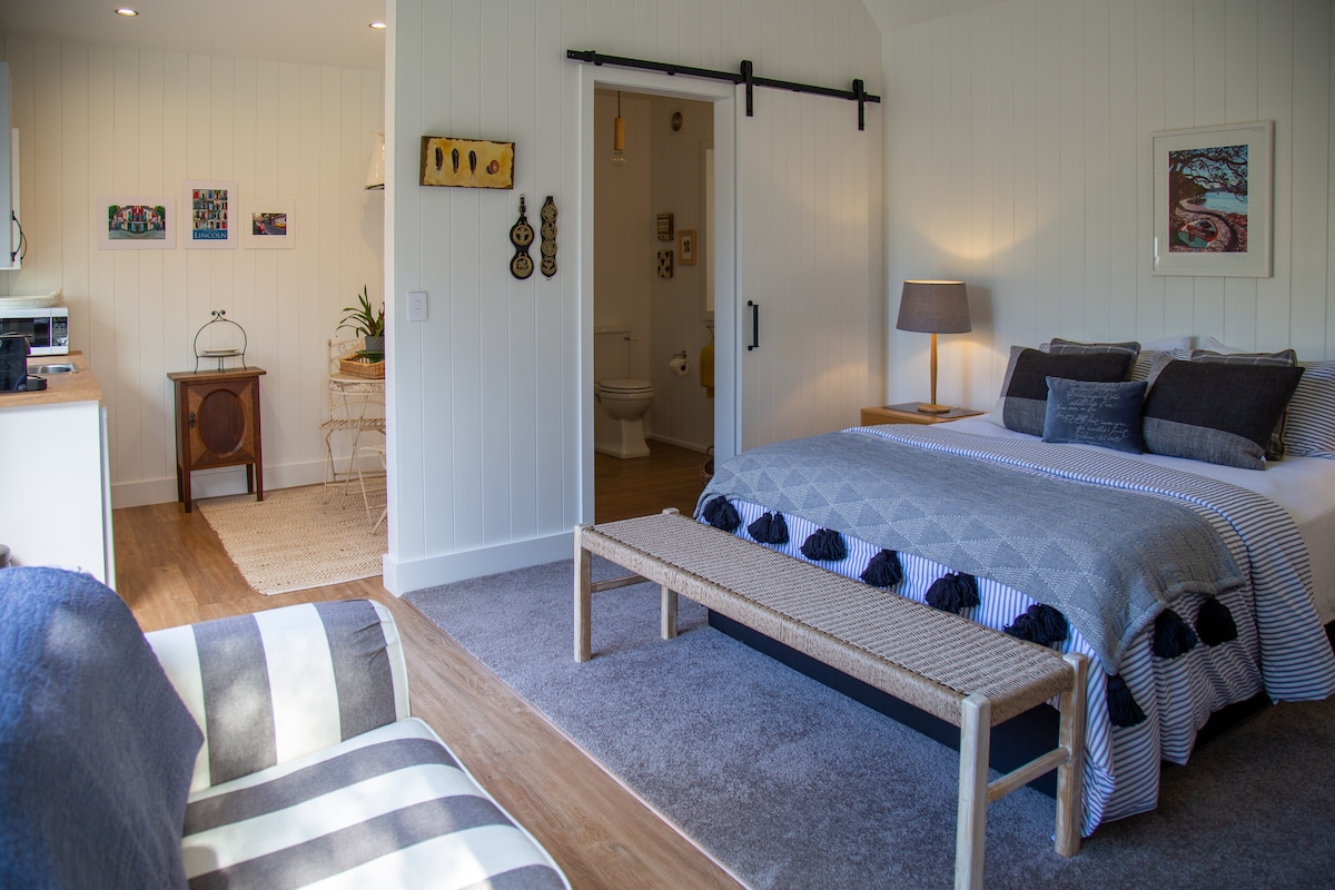 A spacious loft-style cottage interior features a cozy sleeping area with a neatly made bed adorned with gray and blue linens. A comfortable seating area includes a striped sofa, and a sliding barn door leads to an ensuite bathroom, enhancing the open layout.