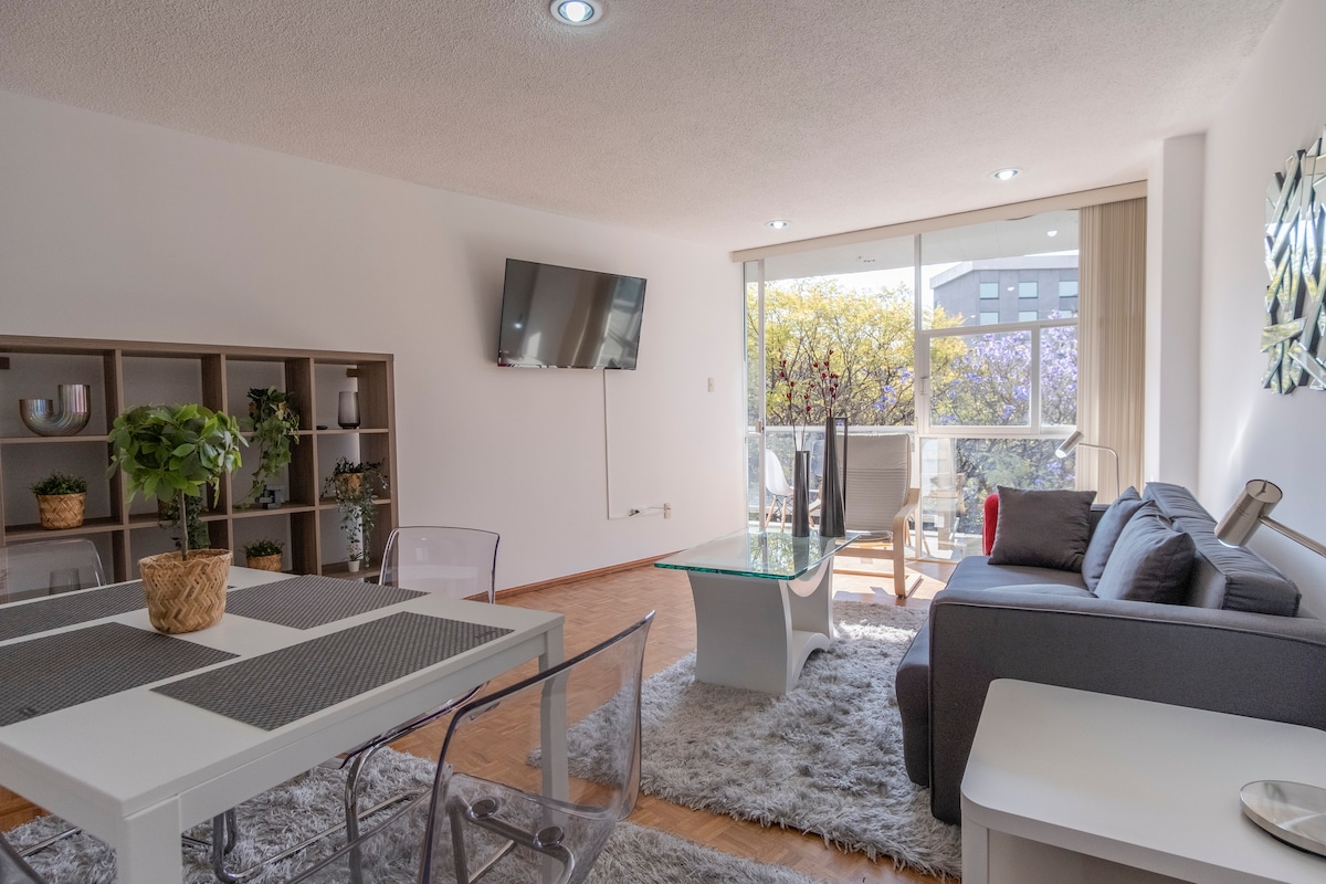 A well-lit living area features a modern gray sofa, a glass coffee table, and a TV mounted on the wall. A dining table with transparent chairs is positioned nearby. Large windows overlook a balcony adorned with blooming trees, providing natural light and a view of the vibrant outdoor scenery.