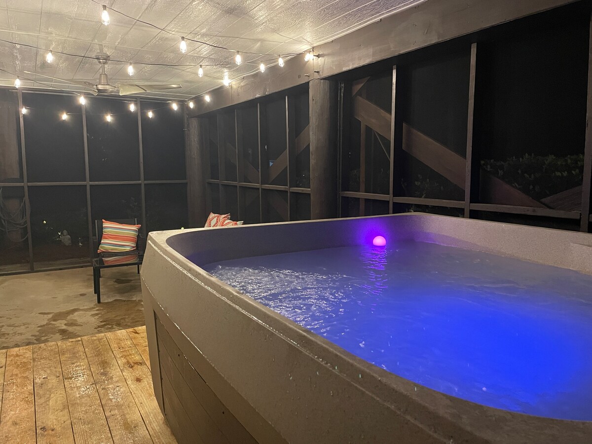 A screened-in patio features a private hot tub with soft, colored lighting. Wooden decking surrounds the tub, and a colorful striped lounge chair is positioned nearby. String lights hang above, creating a relaxed ambiance in the evening setting.