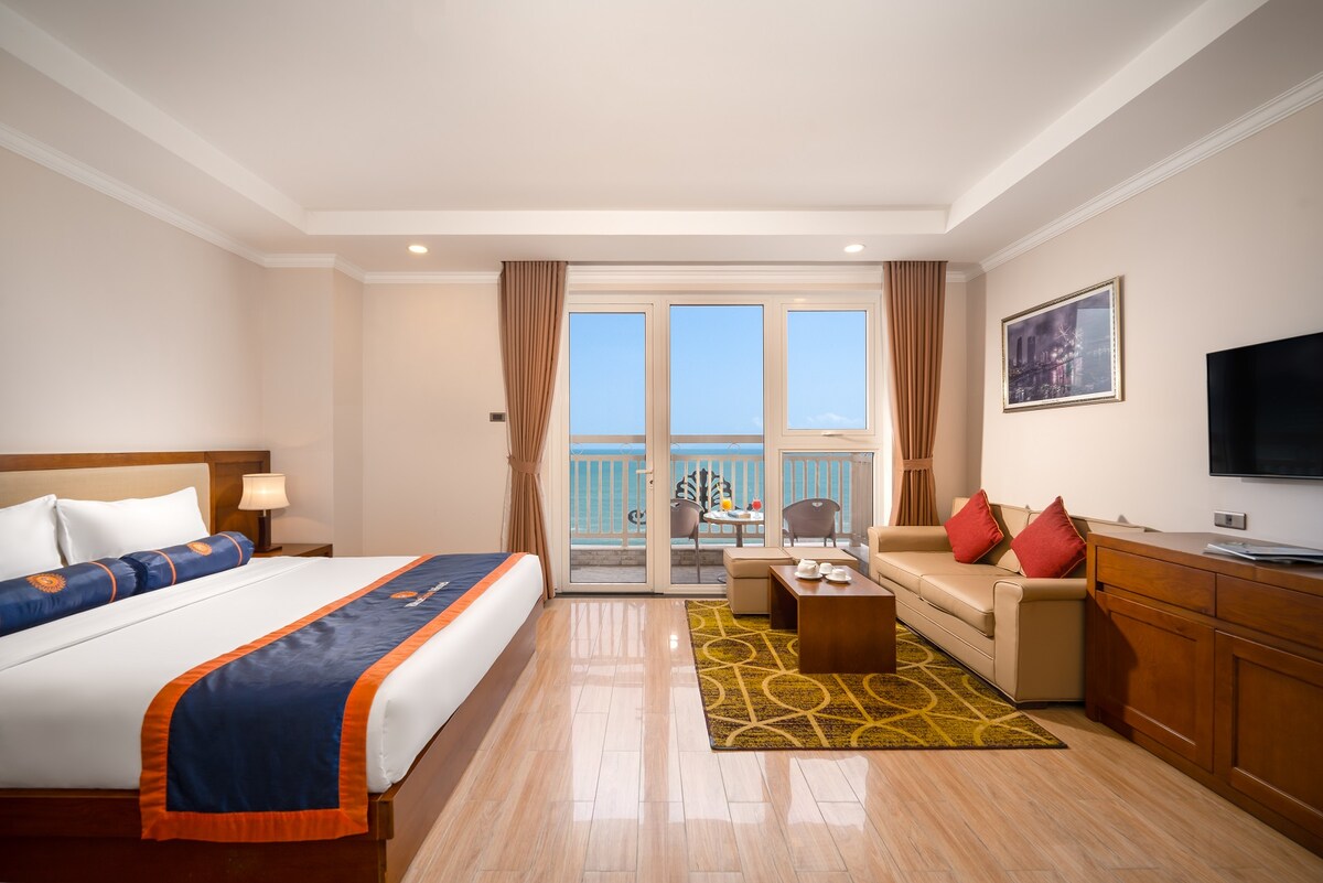 The studio room features a king-size bed with a dark blue and white bedspread. A sofa sits adjacent to floor-to-ceiling windows, revealing a captivating sea view. Natural light fills the space, accentuated by warm wooden furnishings and light-colored walls.