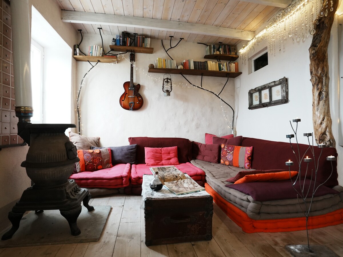 A cozy living area features a large sectional sofa adorned with colorful cushions. A rustic coffee table sits at the center, surrounded by wood accents. Above, shelves display books and decorative pieces, while a guitar is hung on the wall, contributing to a relaxed atmosphere.