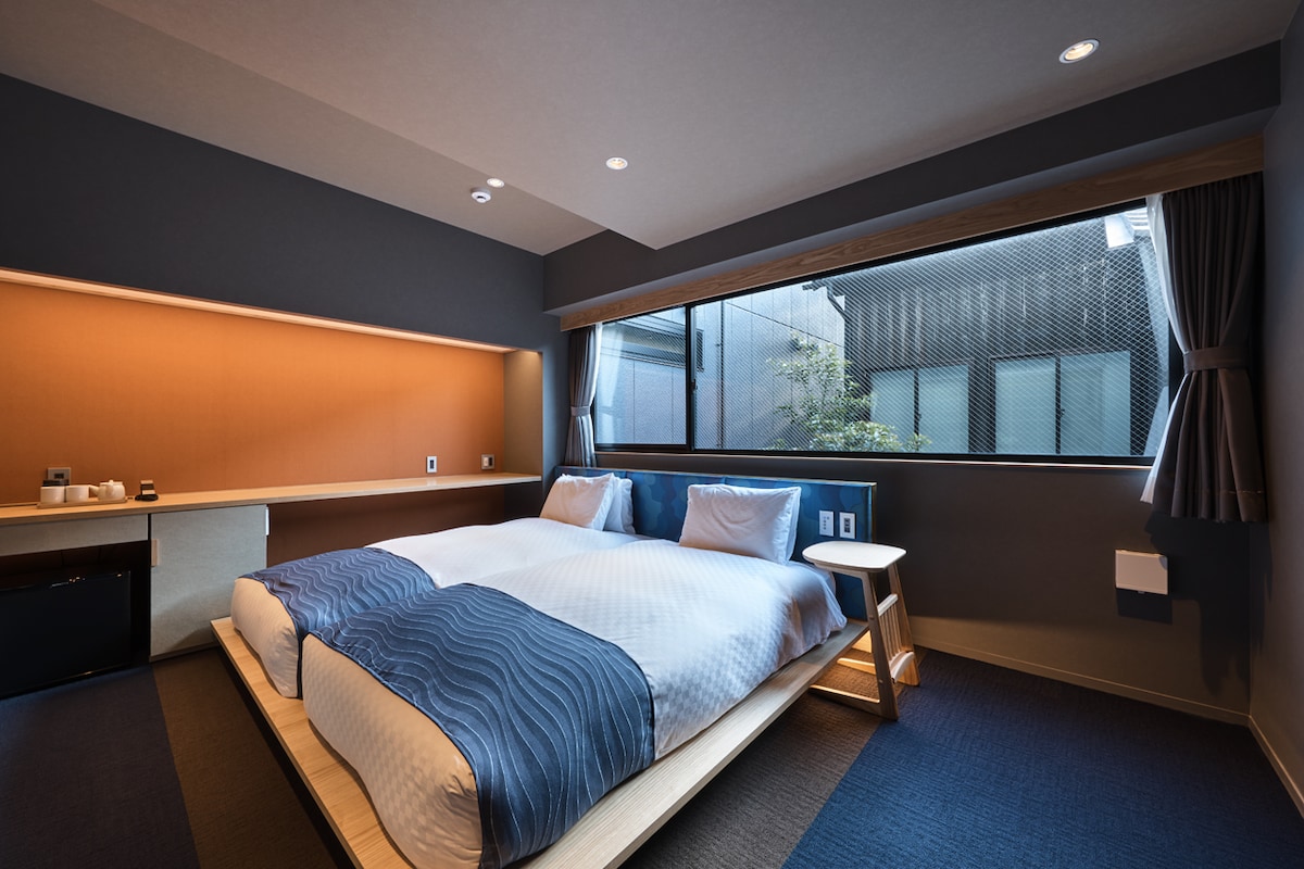 The room is equipped with two single beds, arranged adjacent to each other. A large window provides natural light, offering a view of the outside. A small wooden table is positioned beside the beds, while a minimalist design enhances the overall atmosphere.