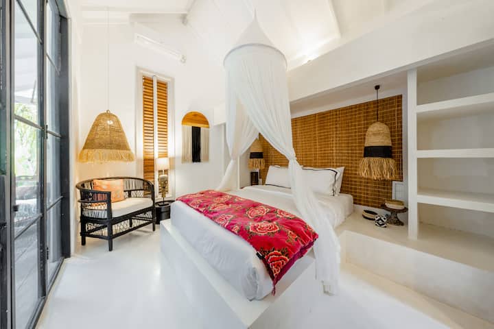 ห้องนอน 3 พร้อมทางเข้าสวน เตียงสี่เสาพร้อมที่นอนคุณภาพสูง ผ้าปูที่นอนและหมอน หัวเตียงทอไม้ ไฟห้อยสไตล์เขตร้อน และเก้าอี้หวาย เปิดประตูกระจกบานใหญ่เข้าสู่สวน สว่างและสงบ