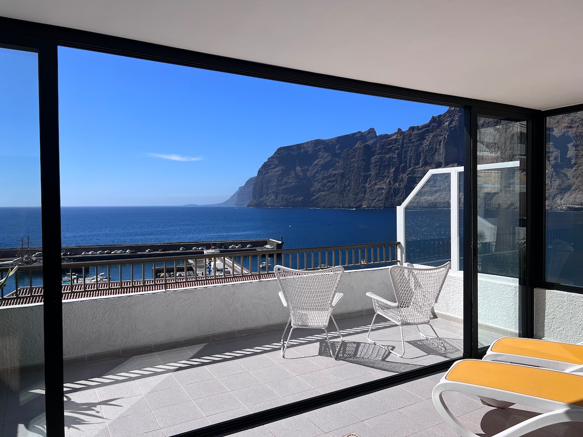 A spacious terrace is framed by floor-to-ceiling glass doors, offering a clear view of the ocean and Los Gigantes cliffs. Two sun loungers are positioned under the sun, creating an inviting space for relaxation and enjoyment of the surrounding scenery.