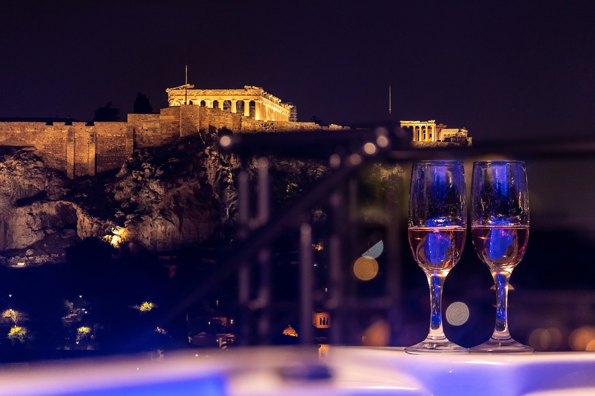 The image features two glasses filled with rosé wine, positioned in front of a view of the Acropolis illuminated at night. The soft lighting accentuates the historic structure against the dark sky, while subtle reflections shimmer on the glass surfaces.