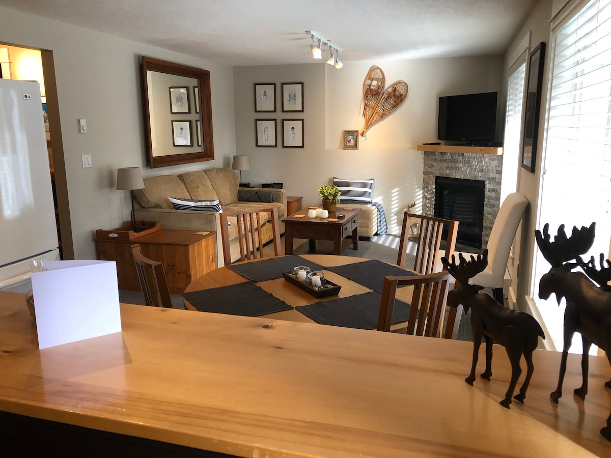A welcoming living space features a comfortable sofa and a rustic coffee table, complemented by framed art on the walls. Natural light enters through large windows, illuminating a dining area with wooden chairs. A fireplace enhances the cozy atmosphere, with decorative accents present.