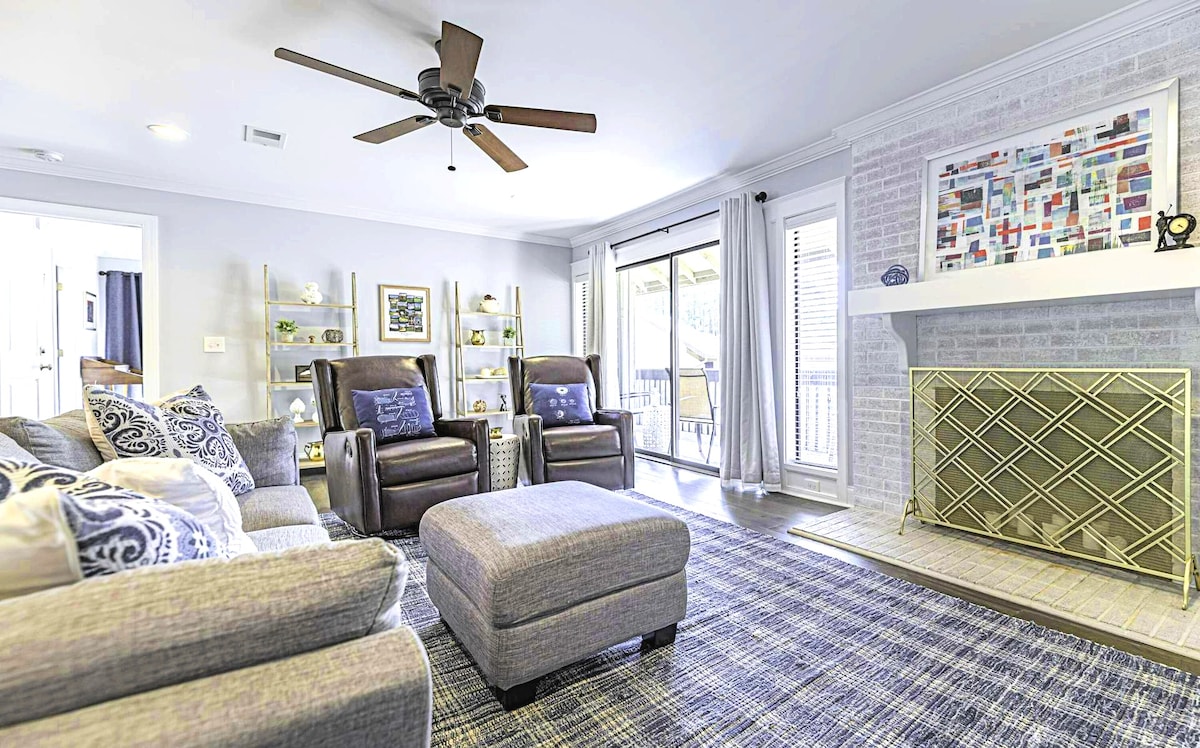 A comfortable living area displays a sectional sofa with decorative pillows beside two armchairs. A ceiling fan hangs above, and a gas fireplace with a patterned front is present. Large glass doors lead to an outdoor space, with a textured rug anchoring the arrangement.