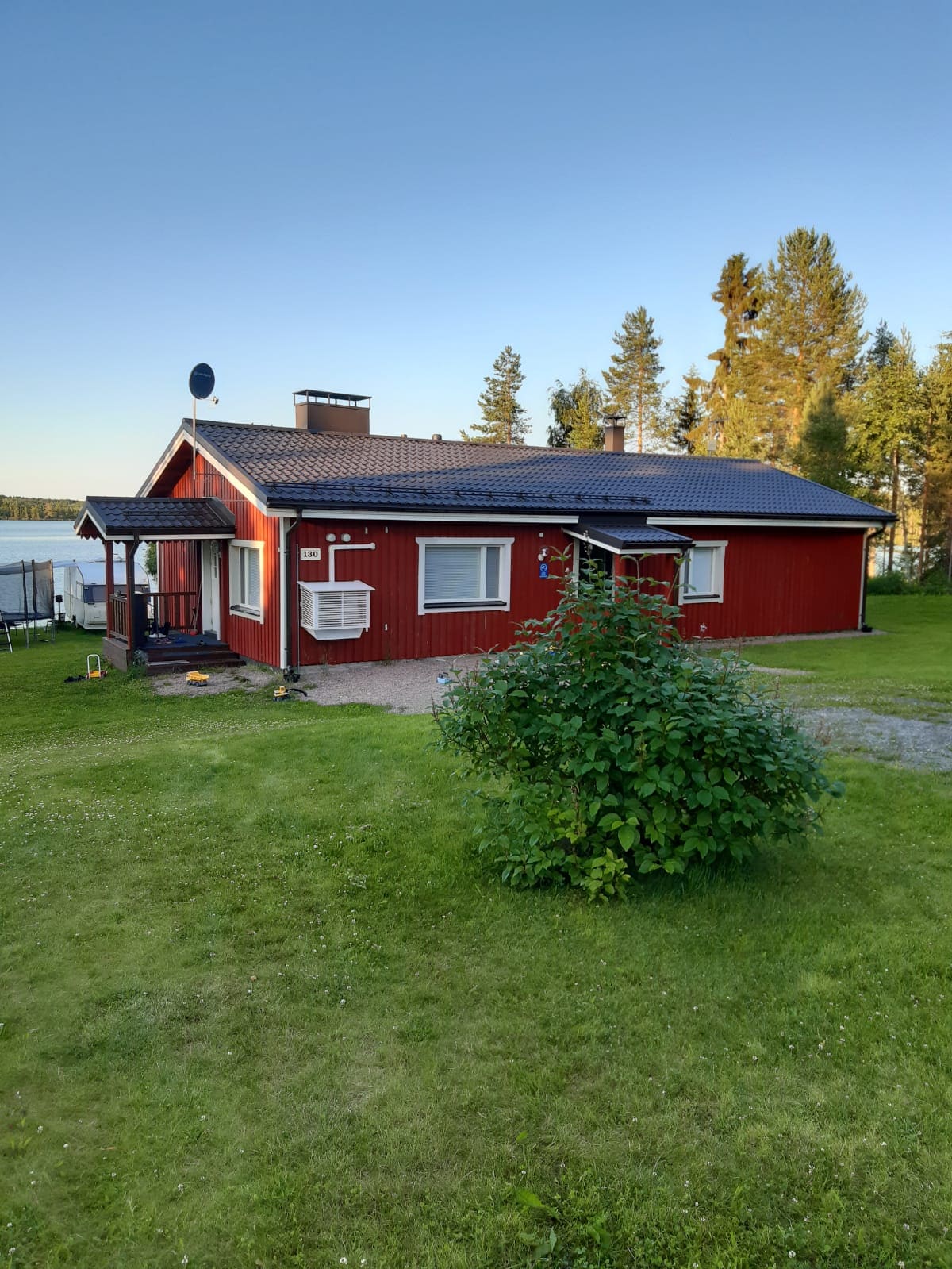 A red wooden house is positioned on a grassy lot by the lake. The exterior is complemented by a covered porch and a satellite dish on the roof. Tall trees frame the background, and a clear sky enhances the serene atmosphere of the surroundings.