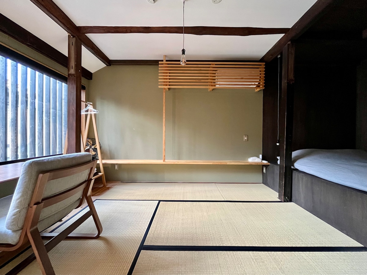 La chambre est un style japonais moderne avec un sol en tatami et des murs plâtrés, la climatisation et un lit Queen Size dans une alcôve en bois foncé. 