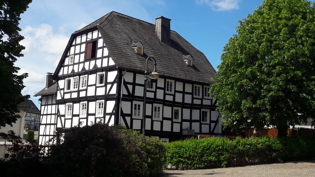 A traditional half-timbered building is showcased, featuring a distinctive black and white facade. Large windows are framed within the wooden structure, while lush greenery surrounds the base. A clear blue sky provides a bright backdrop to this charming architecture.