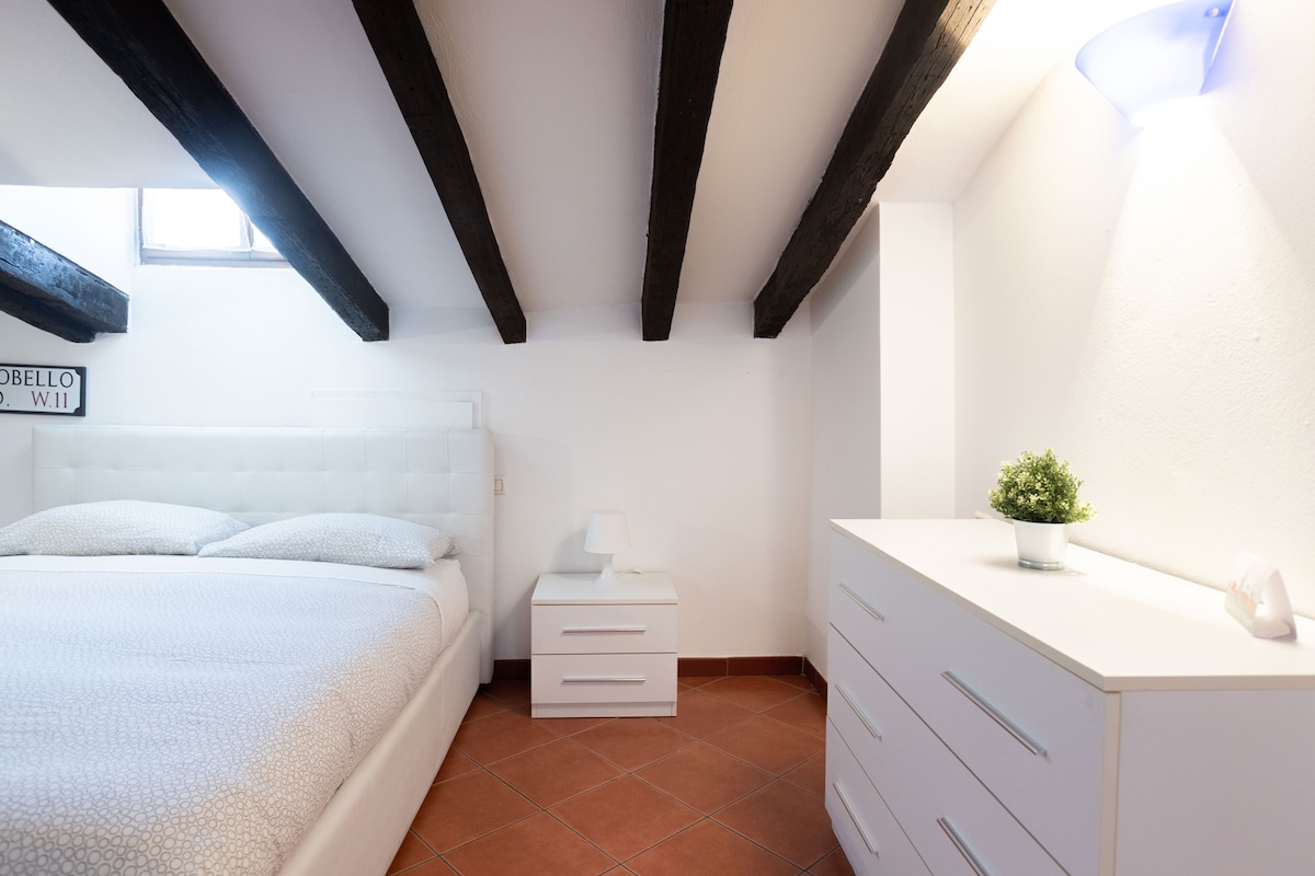 A spacious bedroom features a comfortable bed with a tufted headboard. Light-colored walls are complemented by exposed wooden beams overhead. A white dresser and a bedside table with a small plant enhance the minimalist decor. Natural light enters through a window, illuminating the room.