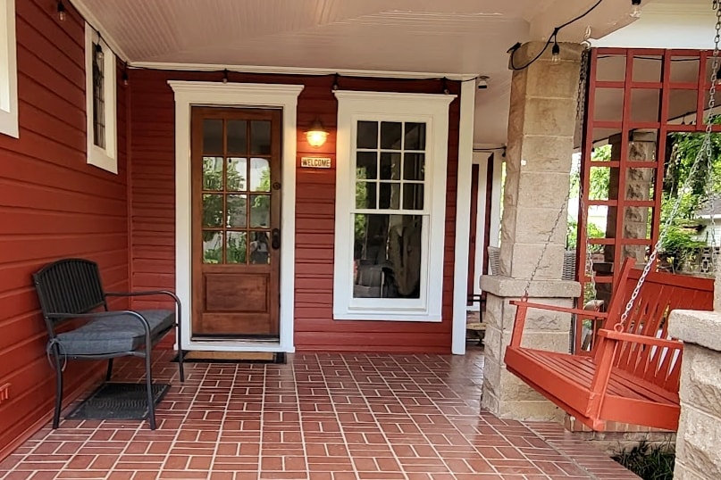 The entrance features a welcoming porch with a wooden swing and seating area. A front door with glass panels is framed by multiple windows, allowing natural light to fill the space. The terracotta tiled floor enhances the inviting atmosphere.