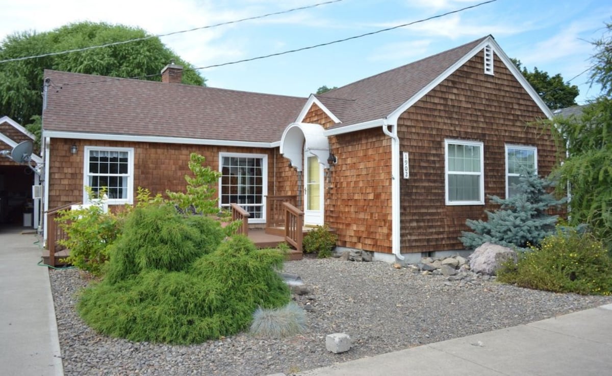 The exterior of the home features a warm wood shingle facade, complemented by large windows. A welcoming front porch with steps leads to the entrance, surrounded by landscaped greenery and decorative stones.