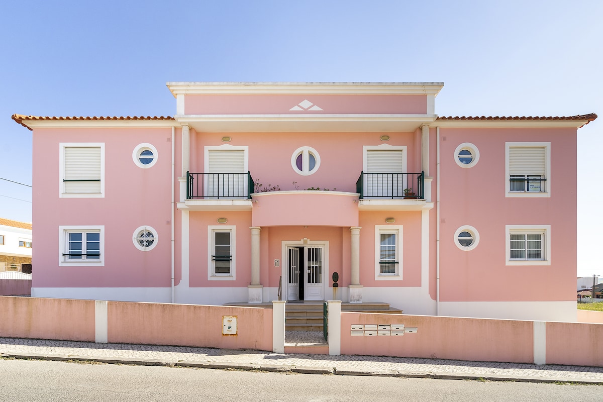 A two-story building features a light pink facade, complemented by white circular and rectangular windows. The entrance is framed by two balconies and decorative columns, leading to steps that provide access to the main door. The structure stands against a clear blue sky.