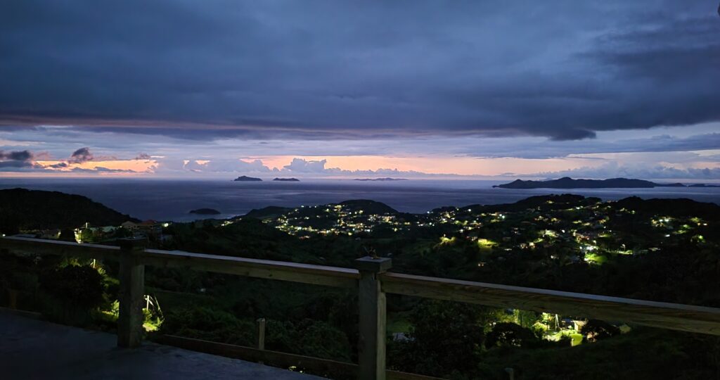 A panoramic view is presented, showcasing the evening sky transitioning from dusk to night, with illuminated coastal areas and distant islands. The scenery is framed by a wooden railing, emphasizing the tranquil landscape and sparkling lights below.