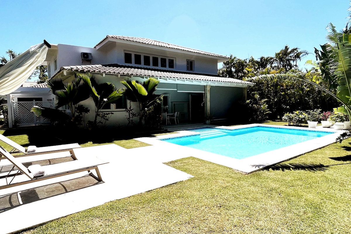 A spacious villa is depicted with a clear blue pool surrounded by a well-maintained garden. Sun loungers are arranged on the patio, and tropical plants enhance the serene outdoor space. The two-story structure features large windows that invite natural light.