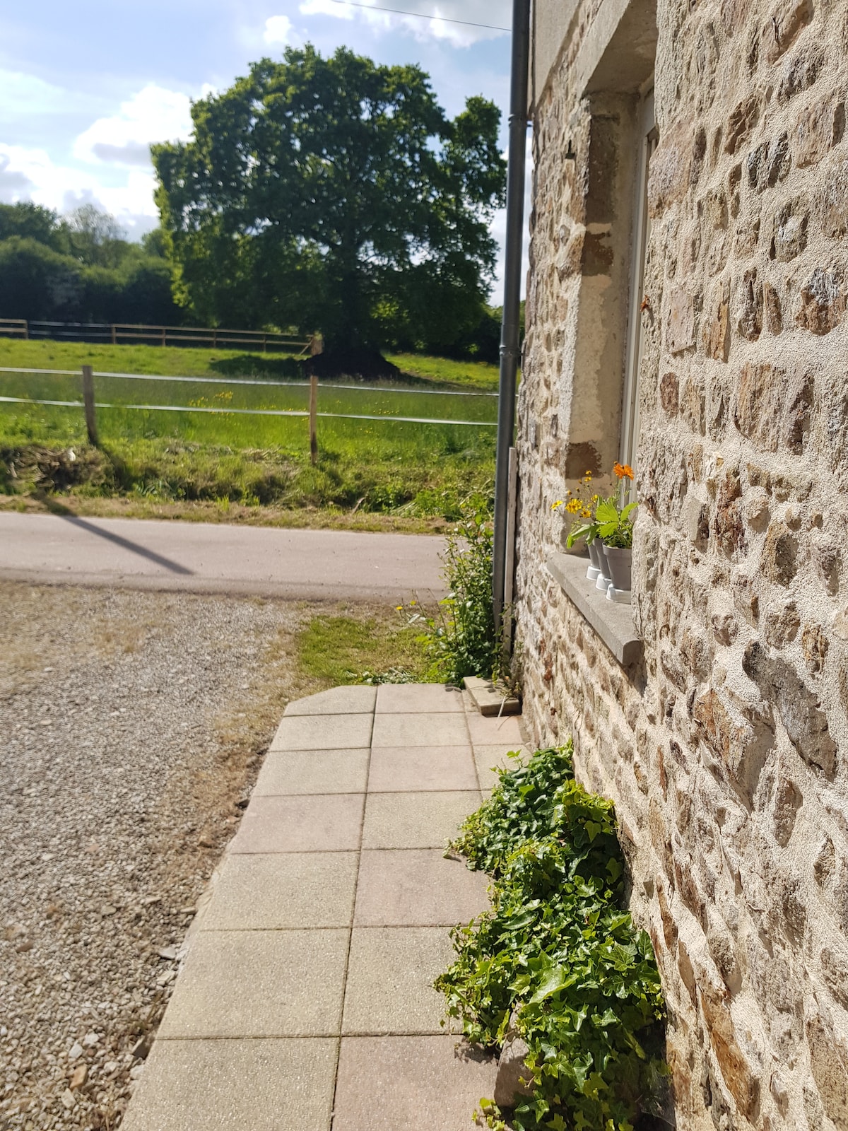 The exterior of the home features a stone façade and a paved walkway. Bright flowers are displayed in a window box, with greenery lining the path. A grassy field and trees are visible in the background, creating a serene countryside view.