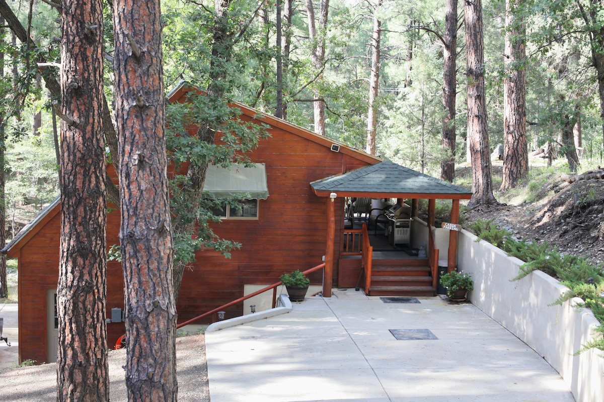 A wooden cabin is nestled among tall trees, featuring a sloped roof and a welcoming porch. Steps lead up to the entrance, surrounded by greenery, providing a serene atmosphere in a natural setting.