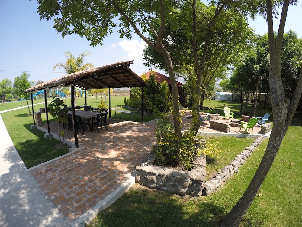 An outdoor area is centered around a covered patio with a wooden roof. Seating options include a long table surrounded by chairs and a fire pit area, complemented by vibrant green trees and neatly trimmed grass. Pathways lead to various spaces, enhancing accessibility.
