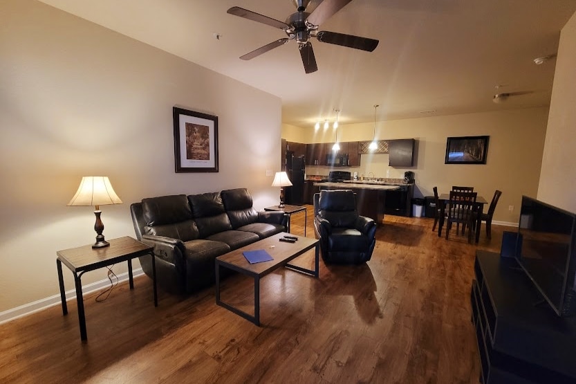 A living area features a dark leather couch and a recliner, complemented by a coffee table. Ambient lighting from a floor lamp enhances the inviting atmosphere. A dining table and chairs are visible in the background, leading to a kitchen with modern finishes.