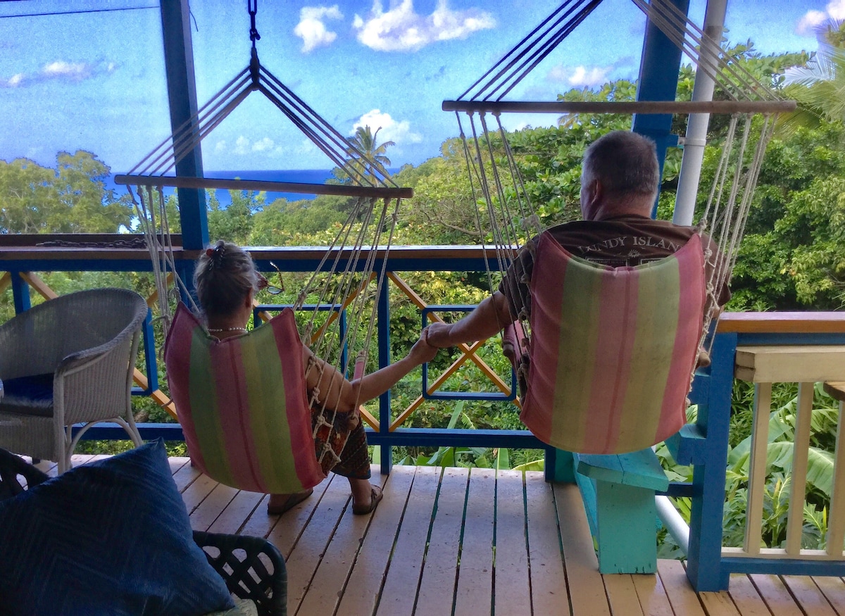 Two hanging chairs are positioned on a balcony, offering a view of lush greenery and the sea in the distance. Natural light filters through the surrounding trees, creating a peaceful setting. The wooden deck is lined with colorful cushions, inviting relaxation.