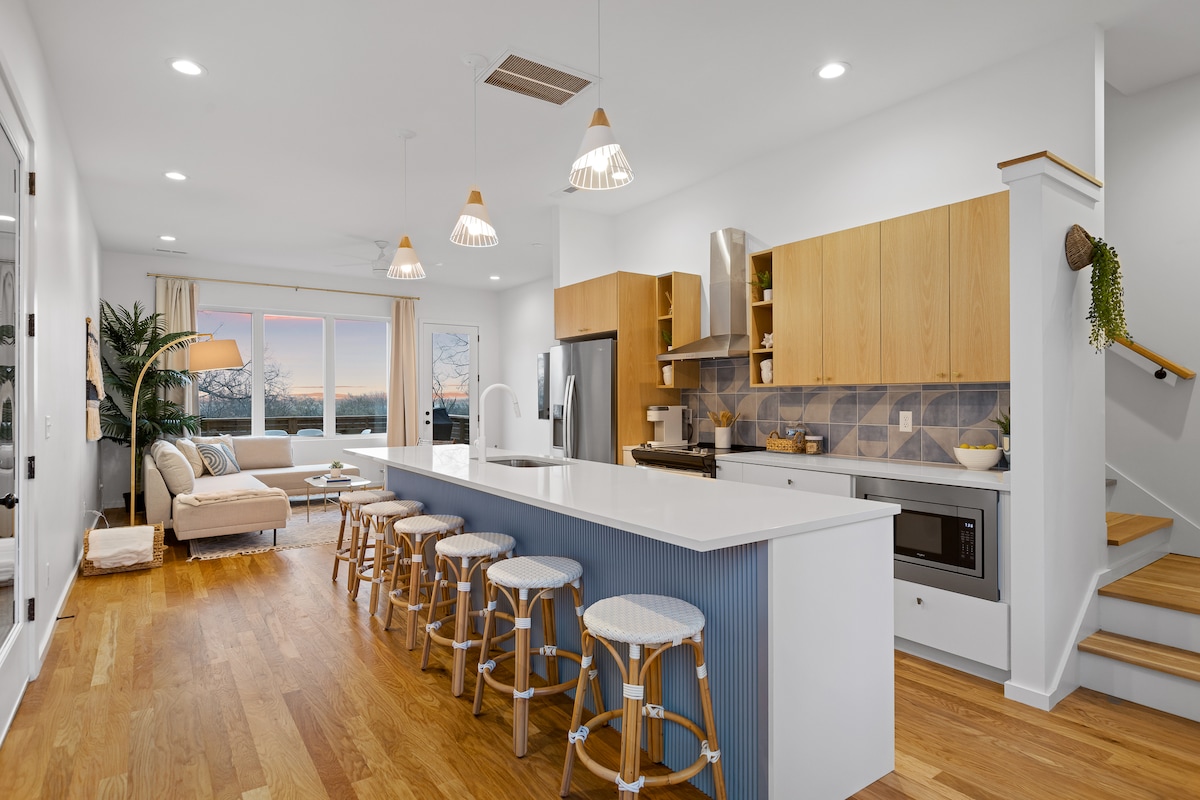 A modern kitchen and living area are featured, showcasing wooden cabinetry and a spacious island seating eight. Light-colored countertops contrast with the patterned backsplash. Natural light floods in through large windows, illuminating the neutral tones and inviting atmosphere of the open layout.