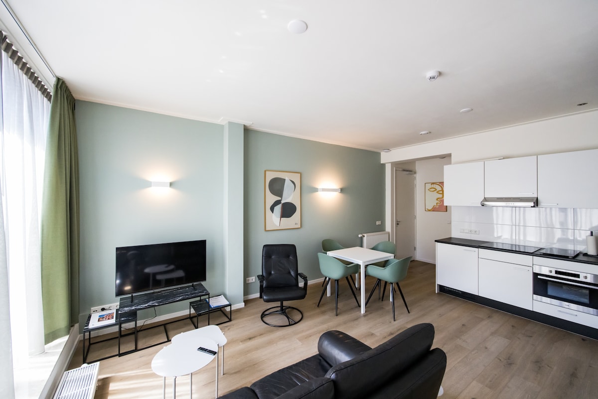 A modern living area is highlighted by a large window with green curtains, allowing natural light to fill the space. A sleek black sofa faces a contemporary television, with a white coffee table nearby. A dining area with a round table and four chairs is positioned across the room.