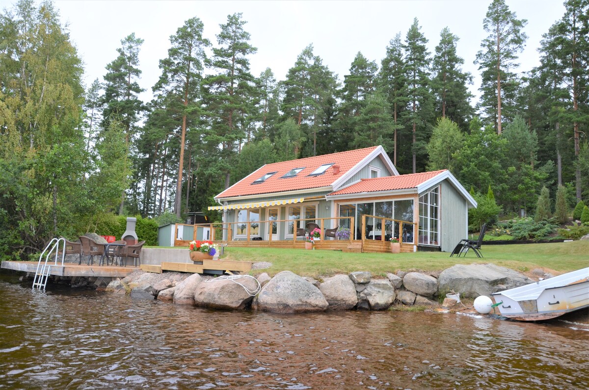 A contemporary house is positioned near the water, surrounded by tall trees. A wooden deck is visible in front, featuring outdoor seating and a ladder leading into the lake. Large windows on the house allow for ample natural light, highlighting its inviting exterior.