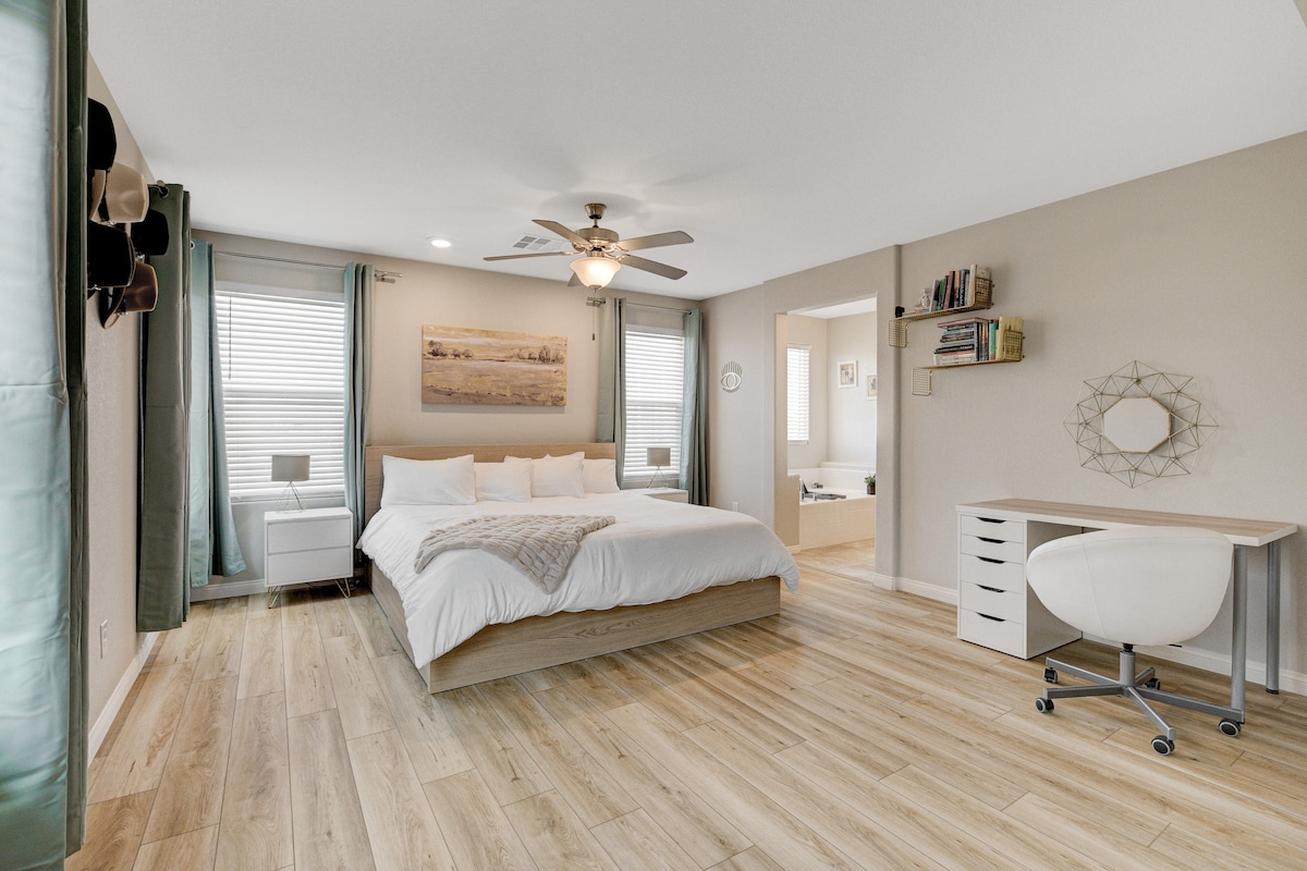 A spacious bedroom features a large bed with a neutral-toned comforter. Two windows with light-filtering curtains allow natural light in. A small nightstand and a ceiling fan add functionality. A desk and chair are positioned nearby, complementing the room's inviting layout.