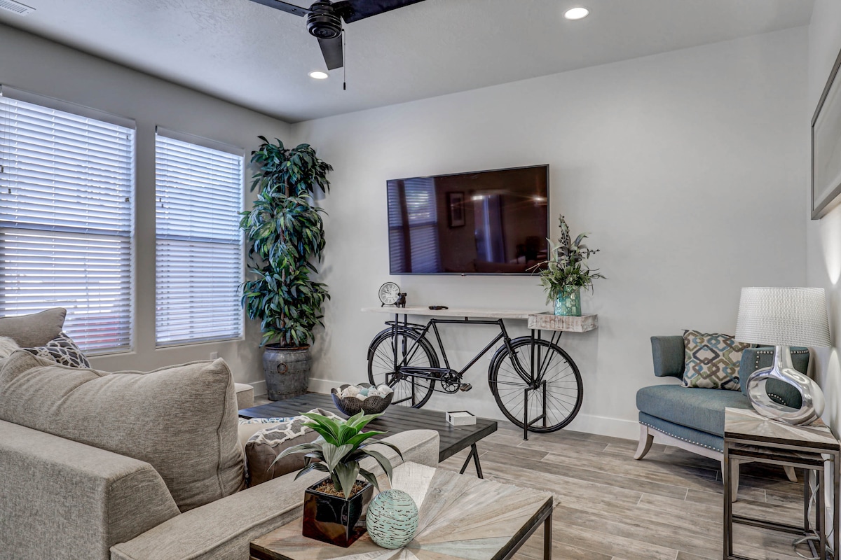 A modern living area features a neutral-colored sofa and a decorative bike mounted on the wall. Natural light streams through multiple windows adorned with blinds. A potted plant adds greenery, and a coffee table showcases tasteful decor, creating a comfortable gathering space.
