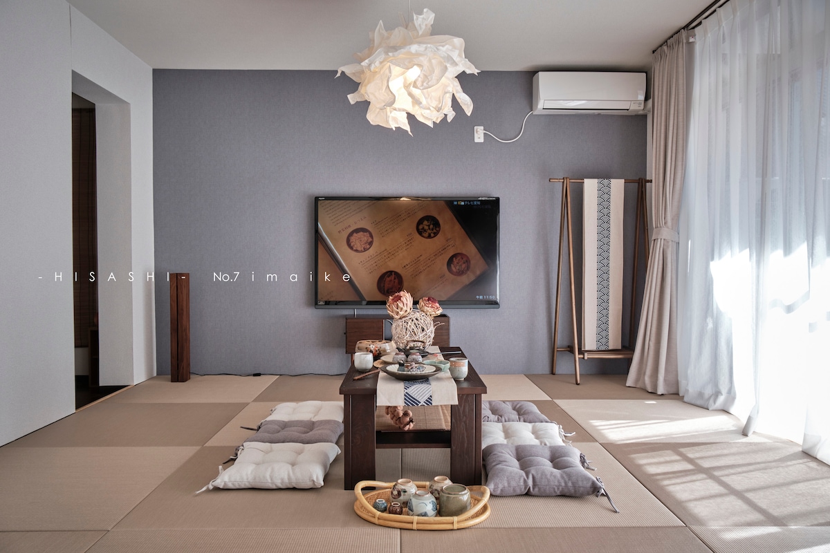 A modern Japanese-style living area is depicted, featuring tatami flooring and a low wooden table. A large television is mounted on the wall, with a delicate paper lamp hanging above. Natural light streams through sheer curtains, illuminating traditional decor on the table.