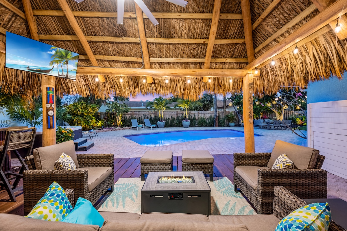 An outdoor seating area under a thatched roof features comfortable lounge furniture arranged around a gas fire table. A flat-screen TV is mounted nearby. The inviting pool is visible in the background, surrounded by tropical landscaping and sun loungers.