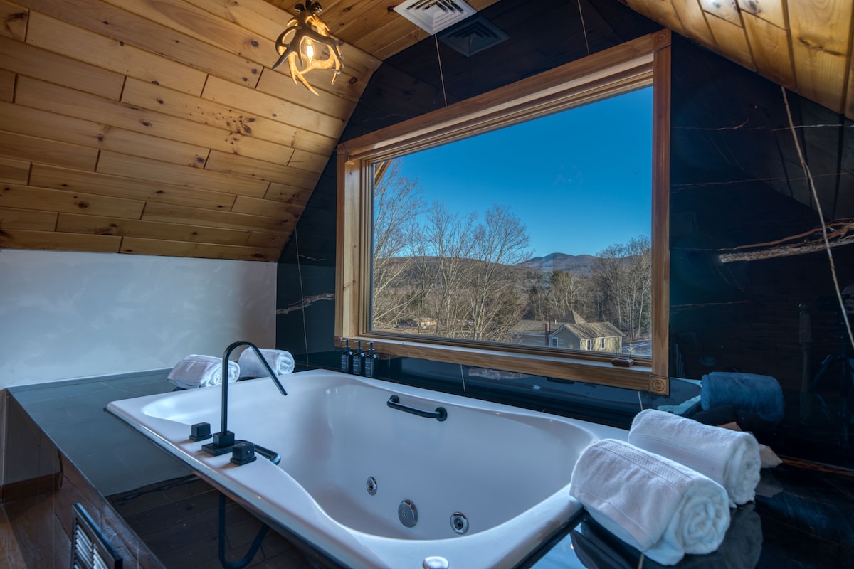 A luxurious marble Jacuzzi tub is positioned beneath a large window, offering scenic views of the surrounding mountains. Plush white towels are neatly rolled and placed nearby, with warm wooden paneling accentuating the elegant ambiance.