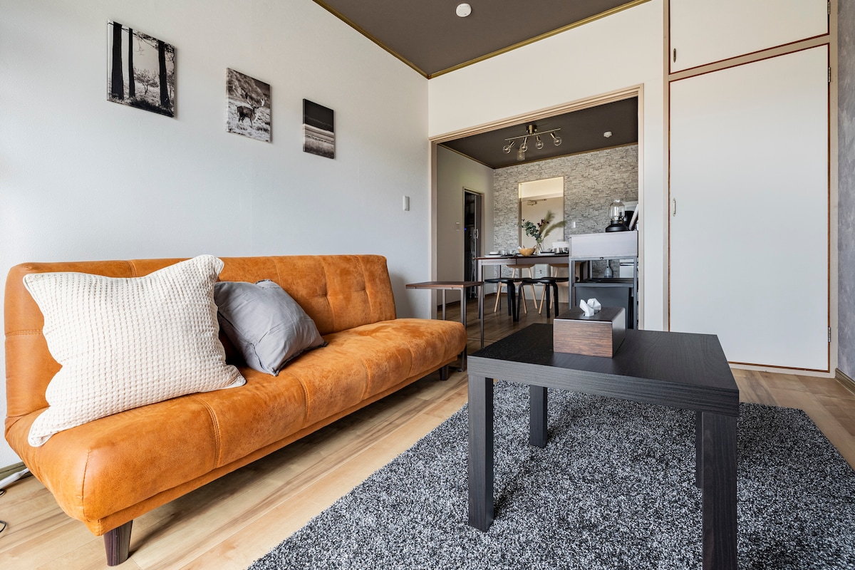 A modern living area features a stylish orange sofa adorned with a textured throw pillow and decorative cushions. A dark coffee table is positioned on a soft rug, with framed photographs displayed on the wall. The space connects to a dining area visible in the background.