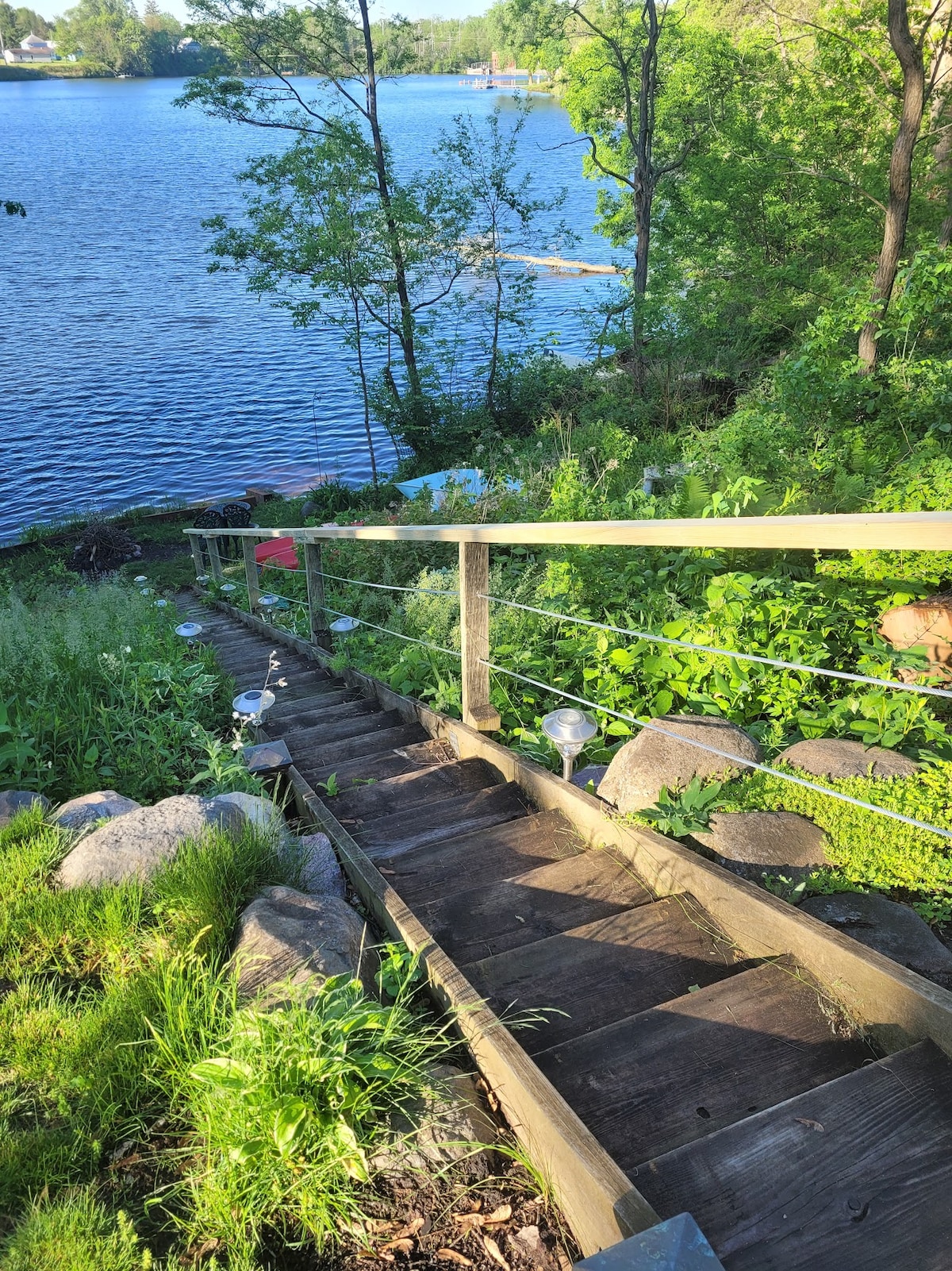 A wooden staircase leads down to the lake, surrounded by lush greenery and rocks. The area features various plants and small lights along the railing, with the serene water visible at the bottom of the steps.