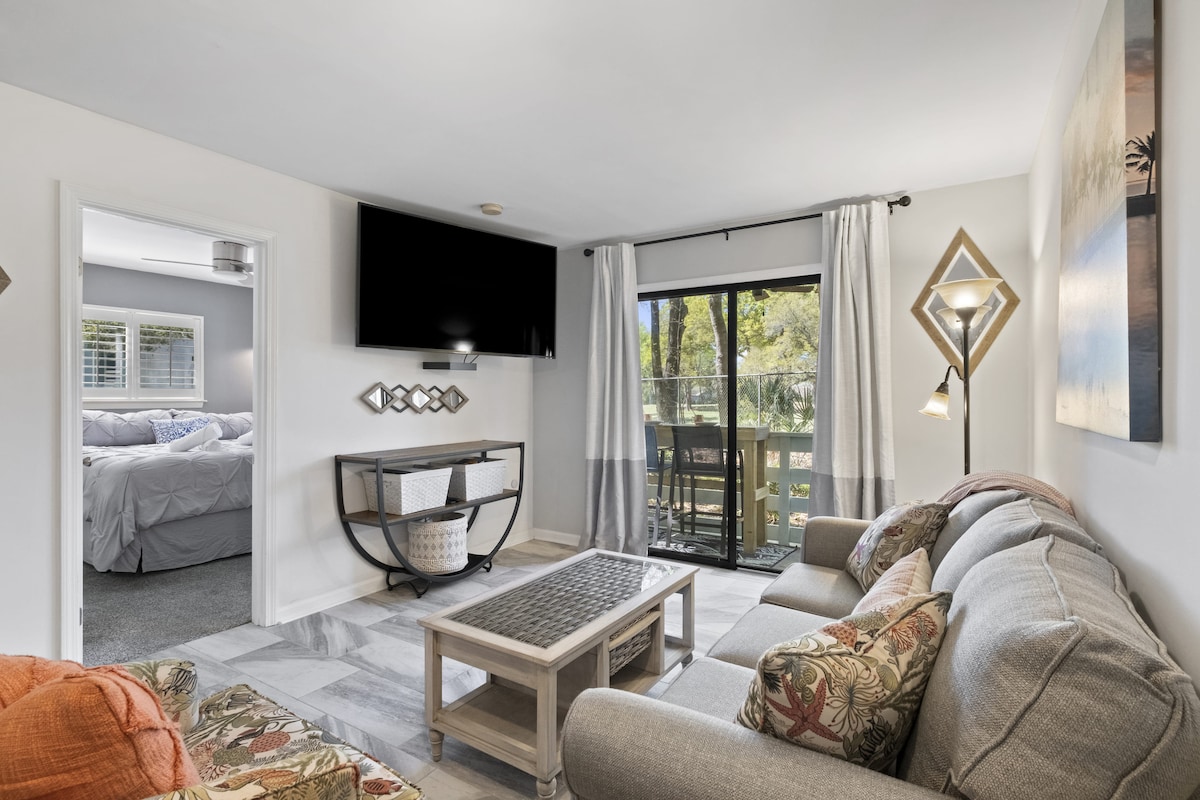 A comfortable living area is highlighted by a spacious couch adorned with decorative pillows. A large flat-screen TV is mounted on the wall, providing entertainment options. Natural light pours in through sliding glass doors, leading to an outdoor deck that overlooks a serene view.