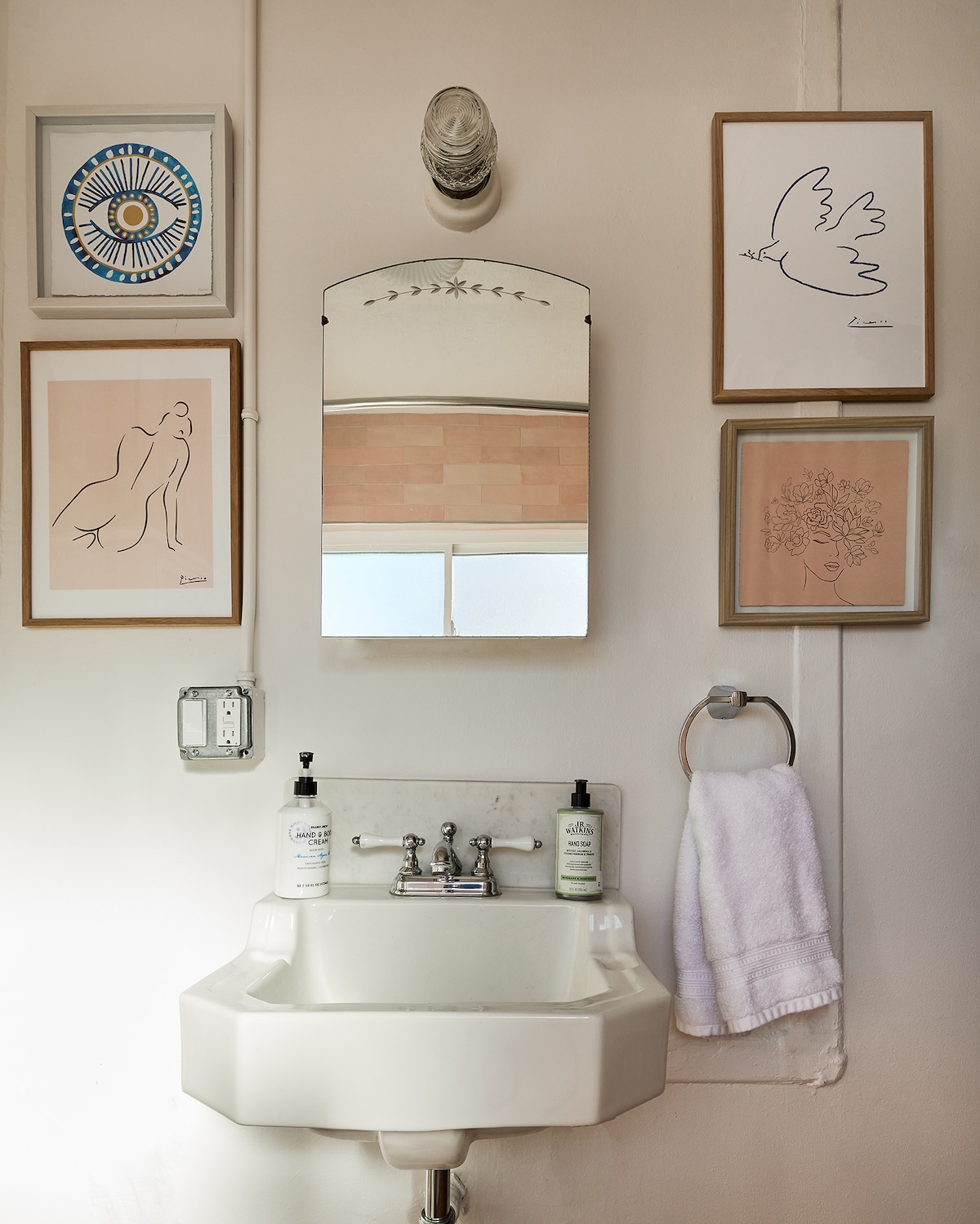 A quaint bathroom area features a white pedestal sink with two chrome faucets. Above the sink, a round mirror is bordered by decorative art pieces showcasing abstract designs. A clean, white towel hangs from a silver ring, adding to the overall simplicity of the space.
