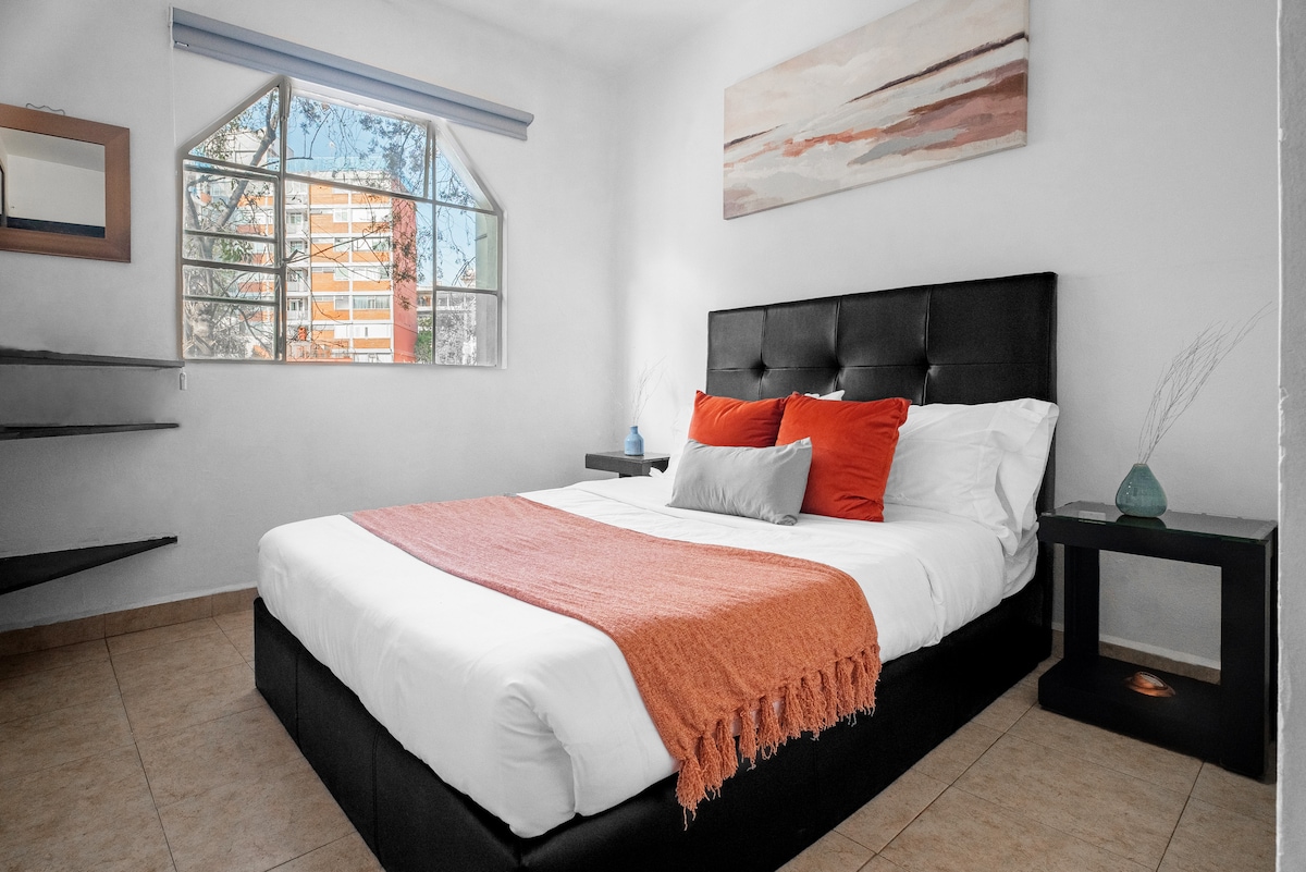 A cozy sleeping area is presented, featuring a spacious bed with white linens and accent pillows in shades of gray and orange. A soft orange throw blanket is draped over the bed. Natural light enters through a large window, illuminating the room's neutral-toned walls and tiled flooring.