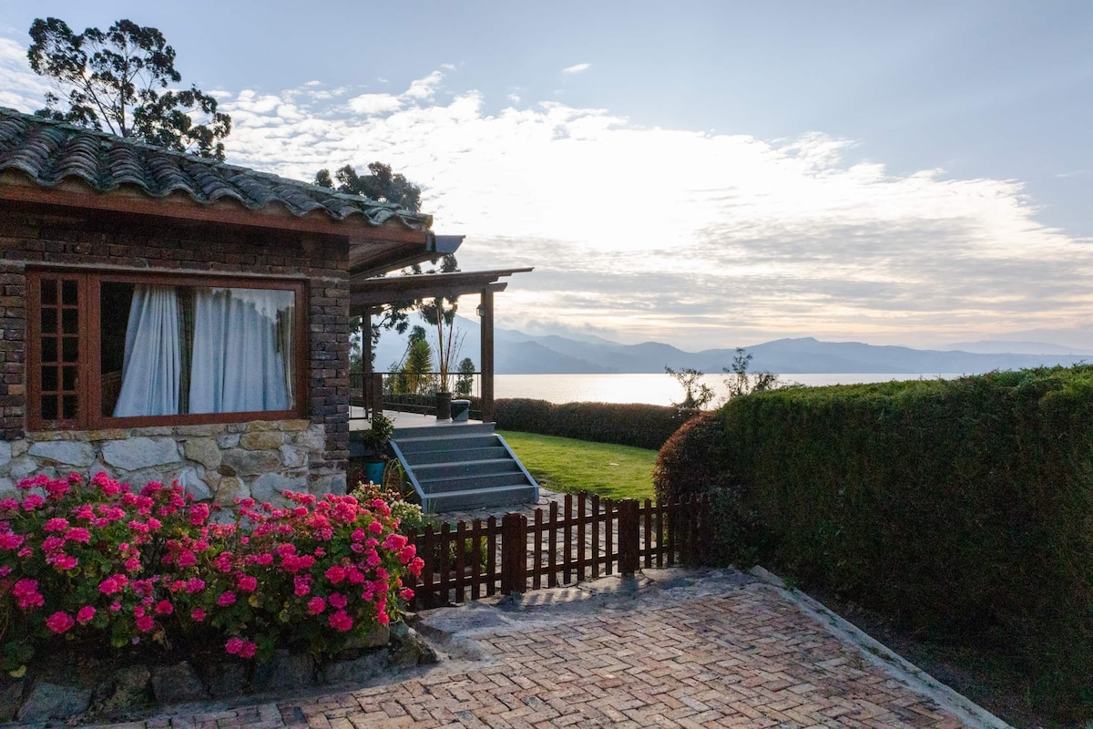 A charming MiniHouse is framed by vibrant pink flowers, with large windows reflecting the natural light. The serene lake is visible in the background, with distant mountains enhancing the scenic view. A wooden fence adds character to the well-maintained outdoor area.